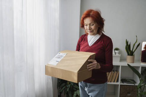Ältere Frau beim Öffnen eines Kartons vor einem weißen Vorhang zu Hause - ALKF01074