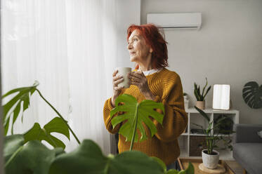 Nachdenkliche ältere Frau mit Teetasse am Fenster stehend zu Hause - ALKF01084