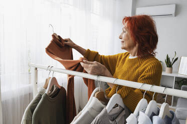 Retired redhead senior woman choosing sweater from clothes rack at home - ALKF01097