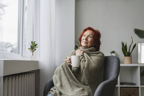 Redhead senior woman sitting in armchair with mug of tea by radiator at home - ALKF01102