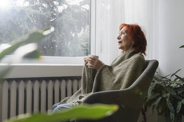 Rothaarige ältere Frau im Ruhestand, eingewickelt in eine Decke, im Sessel sitzend mit einer Tasse Tee am Heizkörper zu Hause - ALKF01104