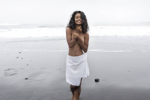 Smiling shirtless woman standing on sand near shore at beach - EAF00143