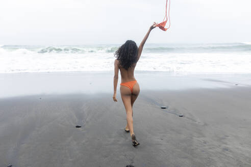 Sorglose Frau, die ihr Bikinioberteil auszieht und auf schwarzem Sand am Strand spazieren geht - EAF00148