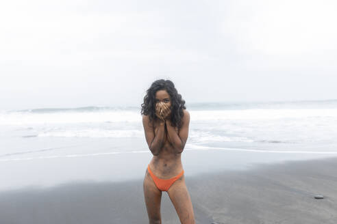 Naked woman covering mouth with hands and standing near shore at beach - EAF00150