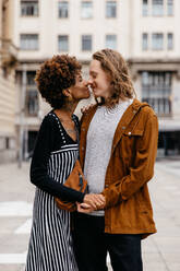 A young couple shares an intimate moment of affection on a city street. Their casual clothing and genuine smiles suggest a natural, authentic connection in an everyday urban environment. - JLPSF31461