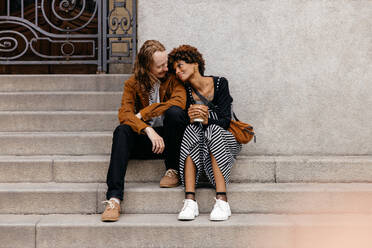 An image capturing a tender moment between a young couple sitting closely on the steps in an urban setting. The candid shot reflects love, companionship, and a casual yet intimate moment shared by two people in love. - JLPSF31469