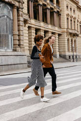 A heartwarming image capturing a young couple in casual wear, hand in hand, crossing the street in an urban setting. The moment reflects companionship and love as they walk together, engaged in conversation. - JLPSF31478