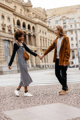 A young and stylish couple enjoys a leisurely walk, hand in hand, along the vibrant city streets, showcasing urban love and connection. - JLPSF31479