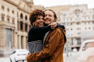 A young couple shares an affectionate embrace on a city street, surrounded by the architecture and hustle of urban life, their joy and connection evident. - JLPSF31483
