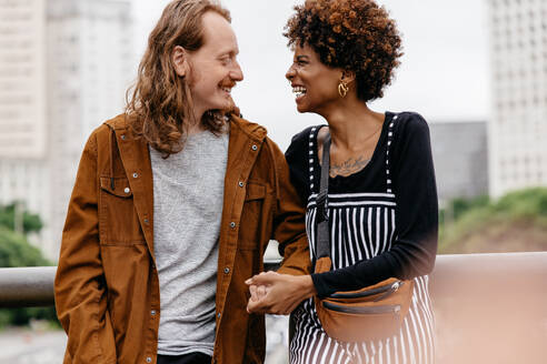 A candid shot capturing the joy and affection of a young couple as they laugh and hold hands during a casual city stroll. Their connection is undeniable in this natural urban setting. - JLPSF31484