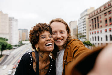 A young couple captures a moment of their urban adventure, taking a selfie with bustling city life and architecture blurred behind them. The image exudes joy, love, and the essence of a modern relationship. - JLPSF31489