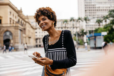 A young and stylish woman standing in an urban landscape, waiting for a taxi in the hustle and bustle of city life. The image captures a woman ordering a taxi via e-hailing on her smartphone. - JLPSF31498