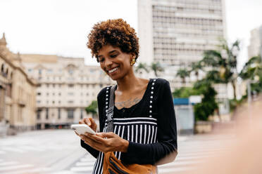 A young woman smiling while looking at her phone, with blurred city street background, captures a moment of urban life and connectivity. Her content expression signifies the joy of an engaging conversation or delightful news. - JLPSF31499