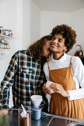 A young, affectionate couple stands in their kitchen, making coffee together. They share a warm, intimate moment as one partner lovingly kisses the other's cheek, creating a cozy, domestic scene. - JLPSF31502