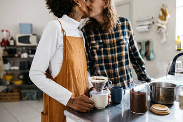 Capturing an intimate moment, a young couple stands in the kitchen surrounded by the warm aroma of fresh coffee. While sharing a kiss, they prepare a comforting cup of coffee together, illustrating a casual, loving domestic scene. - JLPSF31505
