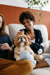 A happy interracial couple enjoys a cozy moment at home with their fluffy small dog. They are seated comfortably on a sofa, sharing affection and smiles in a well-decorated living space. - JLPSF31509