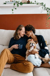 A heartwarming scene of a couple enjoying a quiet moment on a sofa with their adorable dog. They're holding mugs of coffee, exuding comfort and affection in a cozy home environment. - JLPSF31512