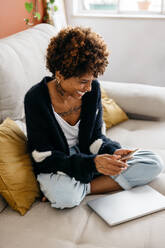 A cheerful young woman with curly hair sits comfortably on a couch at home, casually dressed, looking at her smartphone with a bright smile. A closed laptop lies beside her, indicating a relaxed break from work or study. - JLPSF31518