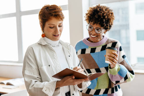 Two colleagues are engaged in a collaborative work discussion in a well-lit freelance office setting. They share ideas and notes in a co-working space, exemplifying teamwork and professionalism in a casual business environment. - JLPSF31530