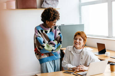 Two cheerful colleagues engage in a collaborative discussion in a bright co-working space. Their casual attire and friendly interaction embody the freelance work environment, surrounded by contemporary office equipment. - JLPSF31544