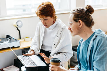 Two professionals engaged in a discussion over a laptop in a well-lit co-working space, exemplifying teamwork and freelance work culture. One holds a coffee cup, suggesting a casual, yet productive, work environment. - JLPSF31567