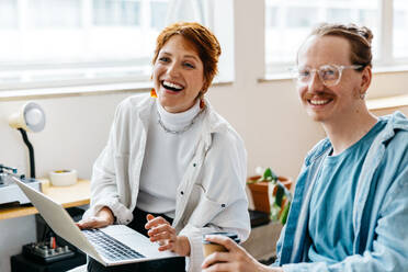 Two happy colleagues sharing a cheerful moment in a modern freelance office setting. They are collaborating in a well-lit co-working space, exemplifying teamwork and positive workplace culture. - JLPSF31569