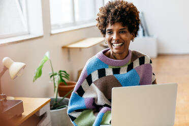 A cheerful young woman with curly hair works on her laptop, sitting at a wooden desk in a bright freelance co-working environment. Trendy, casual attire suggests a creative and relaxed office atmosphere. - JLPSF31576