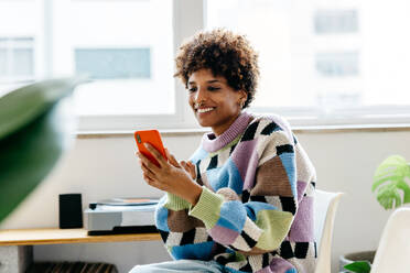 A young female freelancer in a vivid patterned sweater focused on her smartphone while working in a bright co-working office space, conveying a sense of creativity and productivity in a casual work environment. - JLPSF31582