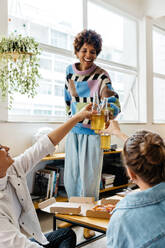 Three colleagues share a cheerful toast with bottles of drinks over a casual meal in a sunlit freelance office setting. A sense of collaboration and relaxation in a modern co-working space is evident. - JLPSF31590