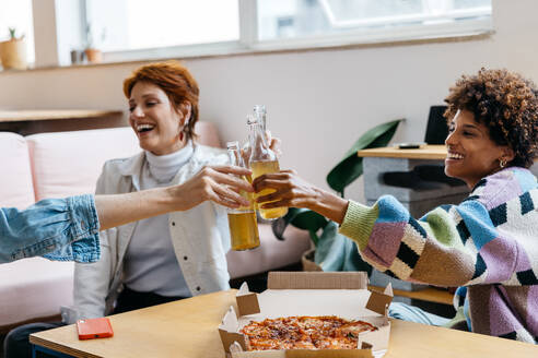 Two cheerful colleagues share a light-hearted moment over pizza and drinks in a modern, sunny co-working space. The informal setting reflects a relaxed freelance office atmosphere, combining work with leisure. - JLPSF31593