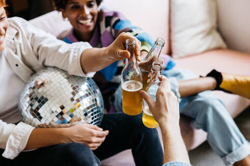 A group of friends casually toasting with beer bottles, enjoying a laid-back party. The mood is cheery with a touch of festivity, as they sit comfortably on a couch with a disco ball suggesting a celebratory atmosphere. - JLPSF31603