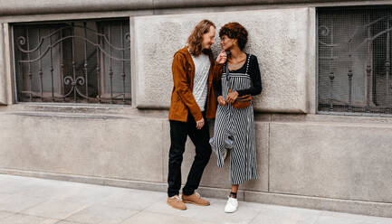 A young couple stands close to each other on a city street, exchanging smiles and enjoying a moment of connection. Their affection is palpable, highlighting a casual urban love story. - JLPSF31617