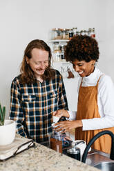 A young, cheerful couple is seen enjoying their time together while making coffee in a cozy home kitchen, surrounded by modern amenities and a hint of greenery. - JLPSF31623