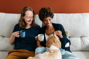 An affectionate moment captured between a happy couple sharing coffee and playing with their pet dog on the couch. The warmth of their home is palpable and invites a sense of joy, relaxation, and companionship. - JLPSF31624