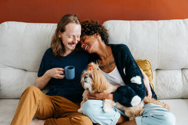 An affectionate couple cuddles with their adorable dog on a sofa, sharing a moment of love and comfort. The man holds a mug, suggesting a relaxed and cozy atmosphere in their home. - JLPSF31625