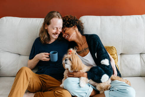 An affectionate couple cuddles with their adorable dog on a sofa, sharing a moment of love and comfort. The man holds a mug, suggesting a relaxed and cozy atmosphere in their home. - JLPSF31625