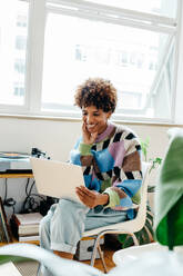 In a brightly lit co-working space, a freelance professional with curly hair is focused and smiling gently while working on her laptop, radiating a sense of productivity and satisfaction in a collaborative office environment. - JLPSF31640