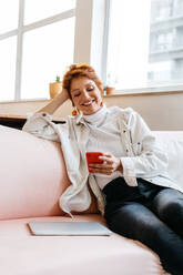 Cheerful young woman sitting comfortably on a pink couch with a smartphone, exuding positivity and ease in a bright freelance office setting. A laptop nearby signifies flexible work. - JLPSF31643