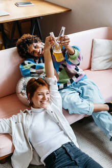 Two happy colleagues enjoy a casual celebration with drinks, lying on a couch in a modern co-working space, exemplifying teamwork and office camaraderie. - JLPSF31653