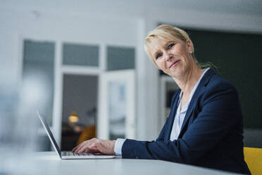 Happy mature businesswoman sitting with laptop at desk - JOSEF23879