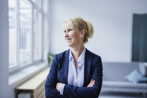 Smiling mature businesswoman standing with arms crossed in office - JOSEF23884