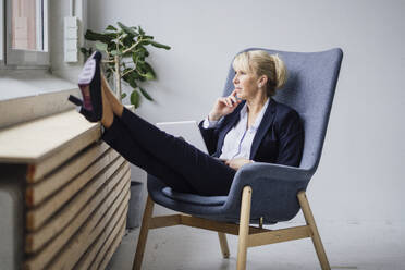 Thoughtful mature businesswoman sitting with laptop on armchair - JOSEF23892