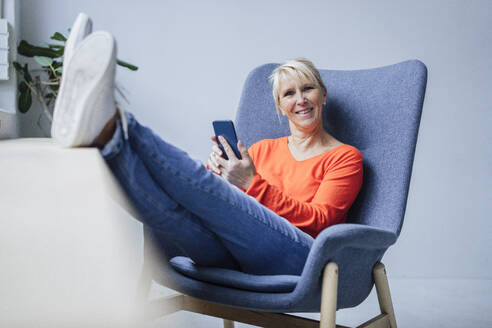 Smiling mature businesswoman sitting with smart phone in armchair - JOSEF23900