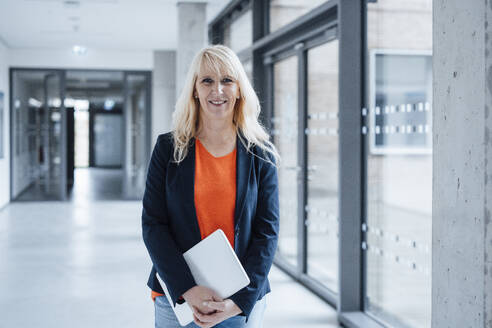 Happy mature businesswoman holding laptop and standing in office corridor - JOSEF23904