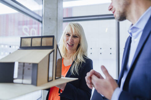 Smiling architect holding model house and discussing with colleague - JOSEF23934