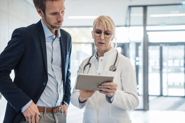 Mature doctor explaining to man over tablet PC in hospital lobby - JOSEF23947