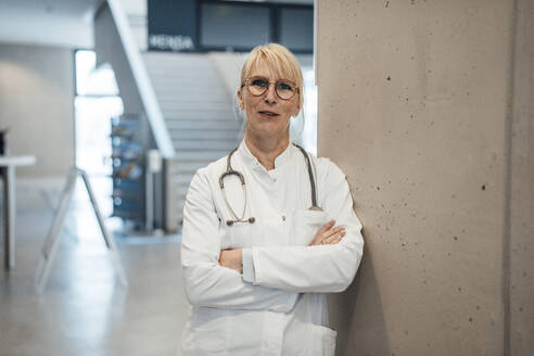 Smiling mature doctor leaning with arms crossed on wall - JOSEF23964