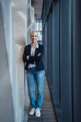 Smiling mature businesswoman standing with arms crossed in corridor - JOSEF23975