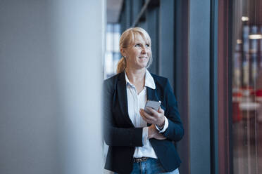 Smiling mature businesswoman standing with mobile phone in corridor - JOSEF23977