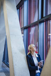 Businesswoman looking up and holding smart phone near columns - JOSEF23978